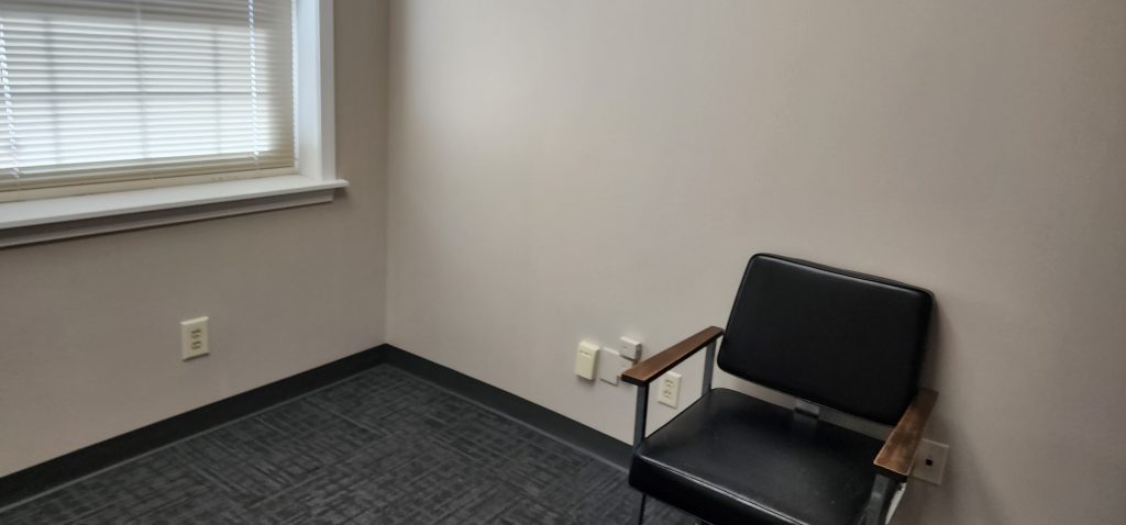Empty office corner with a black leather chair and wooden armrests, next to a window with blinds and beige walls.