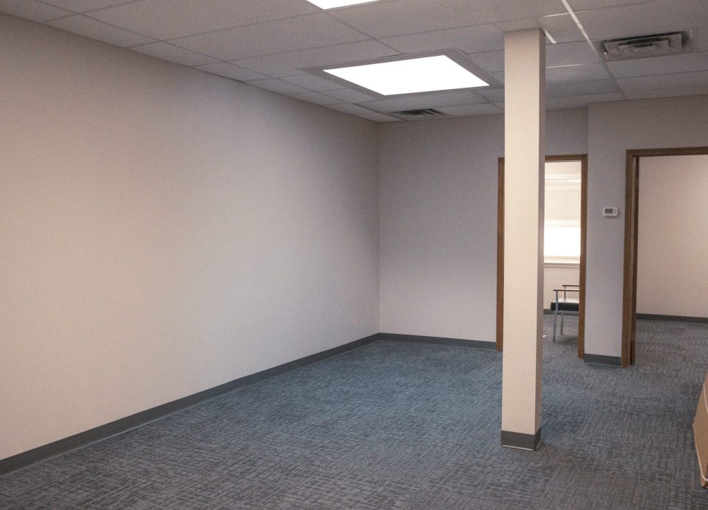 Empty office space with blue patterned carpet, beige walls, and a central pillar, with open doorways to another room in the background.