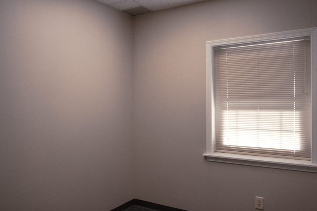 Empty small room with beige walls, dark carpet, and a window with closed horizontal blinds letting in soft light.