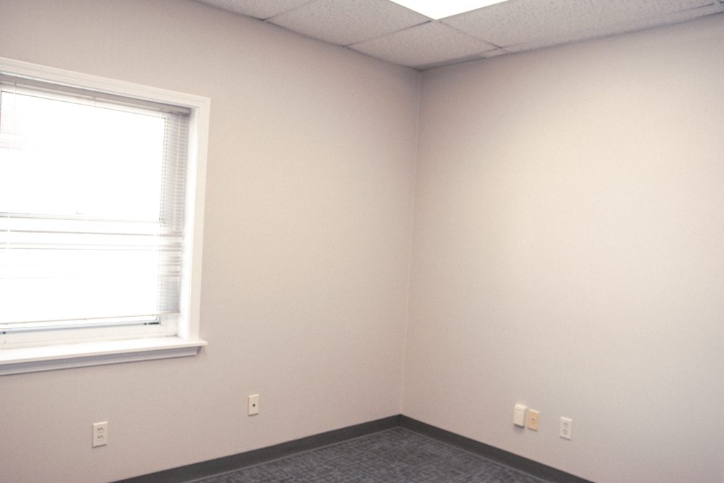 Empty office with light gray walls, a window on the left, and a gray carpeted floor; corner with outlets visible.